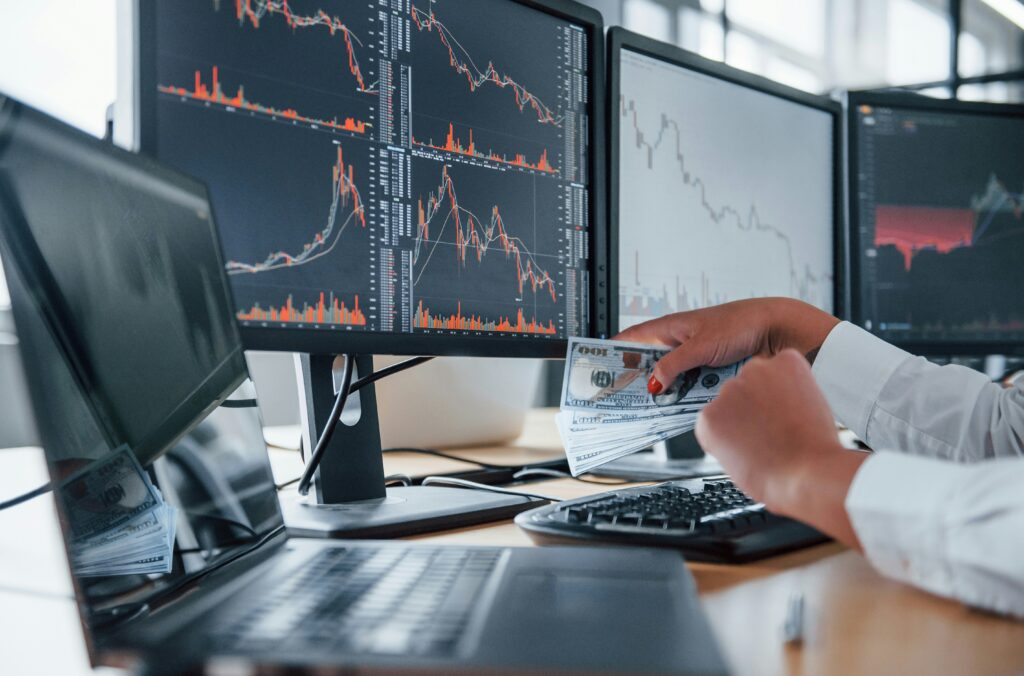 Hands holding money in front of multiple monitors displaying financial charts.