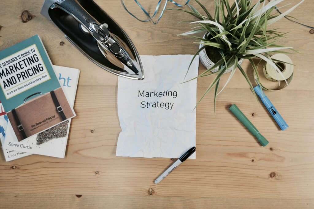 Overhead shot of a book, iron, plant, paper, and pens on a wooden surface. The paper reads “Marketing Strategy”.