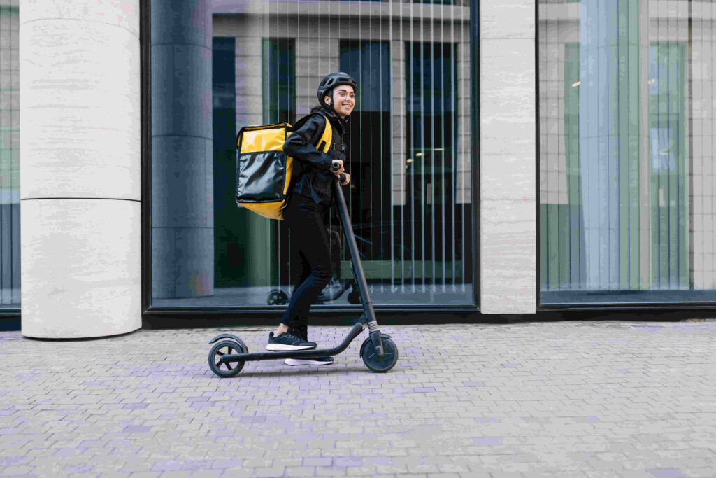 A smiling gig worker in black rides an electric scooter with a large yellow delivery bag on her back in a city.